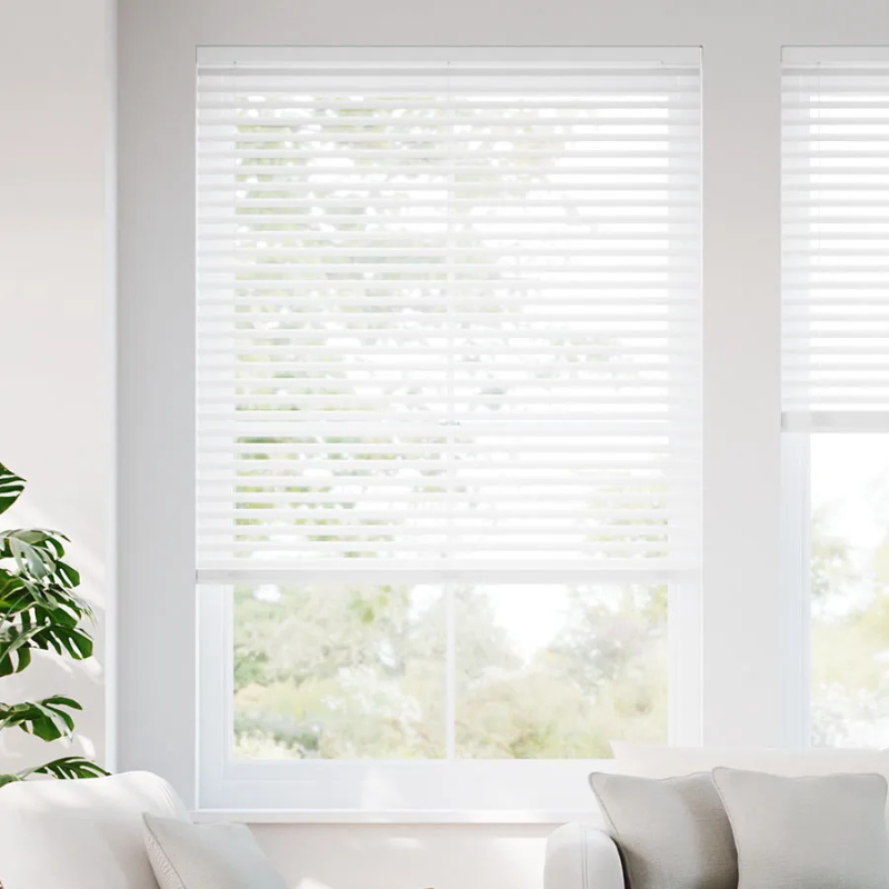 White horizontal window blinds with slats partially open, allowing light to filter through; a plant and a sofa with cushions are nearby.