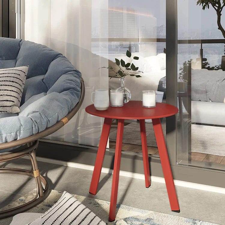 A red round side table with three candles and branches in a vase, accompanied by a wicker chair with blue cushions.