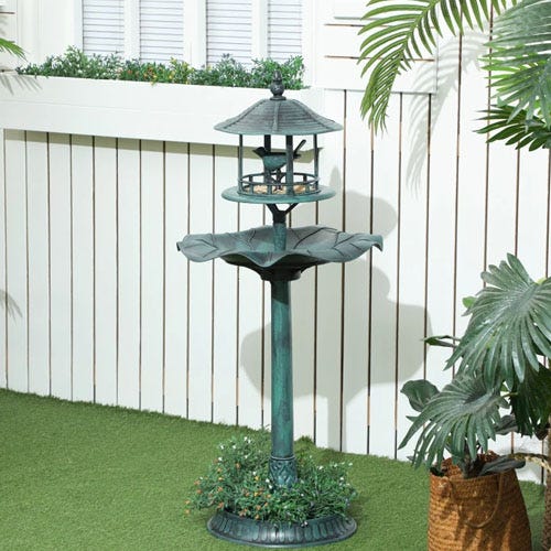 A two-tiered standing bird feeder with a decorative, leaf-shaped basin and a pagoda-style roof is set on artificial grass surrounded by potted plants and a white fence.