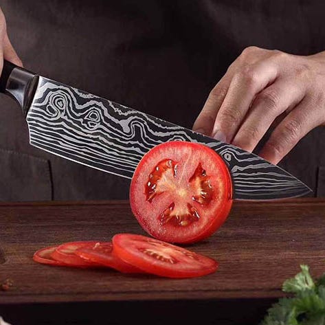 A chef's knife with a patterned blade slicing through a tomato on a cutting board.