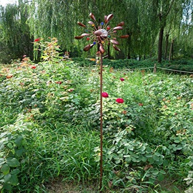 A copper-colored metal wind spinner with dual spinning blades resembling flowers and a solar-powered LED light in the center, stands amidst a garden.