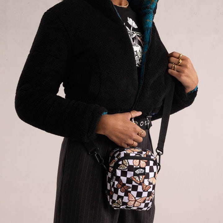 A person wearing a black fuzzy jacket, graphic tee, striped pants, and holding a small patterned shoulder bag with butterflies.