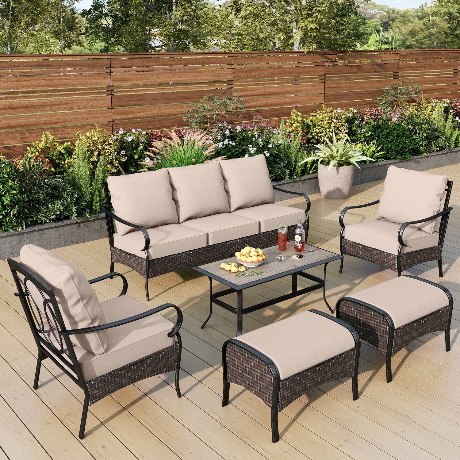 Outdoor furniture set comprising a sofa, two chairs, two ottomans, and a coffee table with cushions and a wooden fence background.