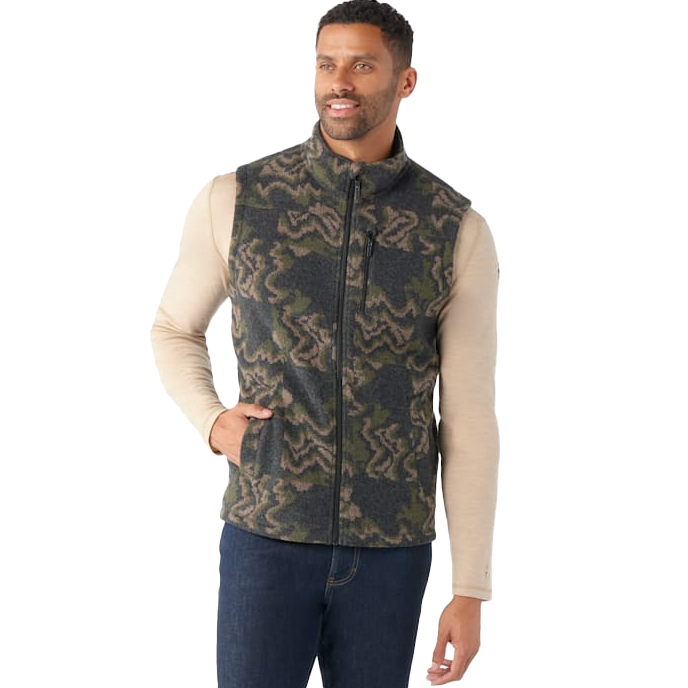 A man wears a Smartwool Hudson Trail Fleece Vest featuring a camo-like pattern in dark green and brown tones. The vest has a full front zipper and is paired with a beige long-sleeve shirt and dark jeans.