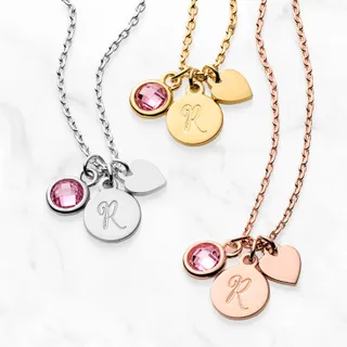 Four pendant necklaces in silver, gold, and rose gold tones, each featuring a heart and a circular charm with the letter 'R' and a pink gemstone.