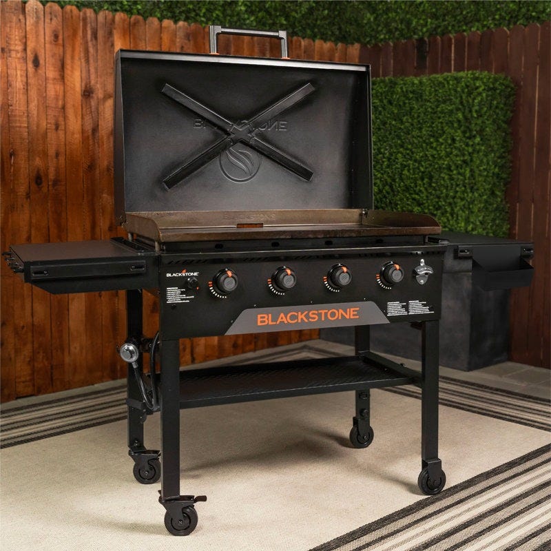 Blackstone outdoor griddle with four control knobs, side shelves, and a hinged lid, suitable for grilling and flat-top cooking. Mounted on a wheeled cart for mobility.