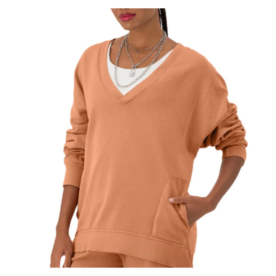 A person wearing a tan V-neck sweater with layered necklaces and pockets.