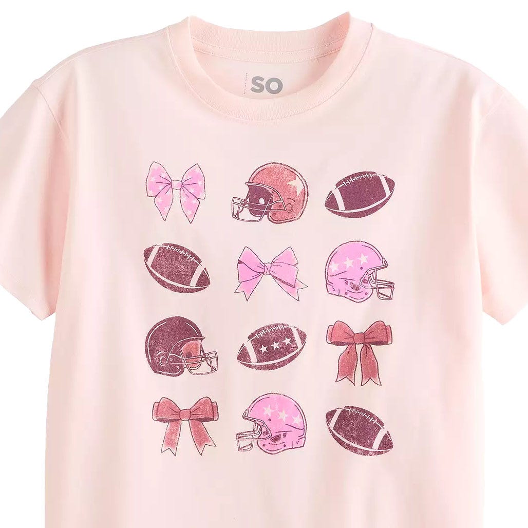 A light pink T-shirt features a pattern of pink and brown football helmets, footballs, and bows arranged in a grid.