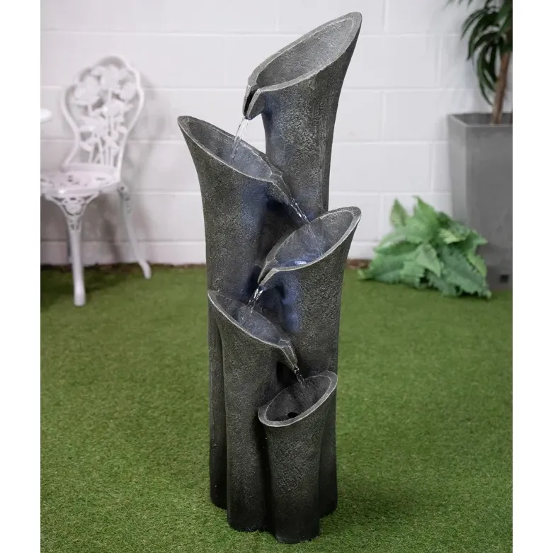 A decorative water fountain with multiple levels, shaped like abstract tubes in grey tones, set against an indoor background with artificial grass.