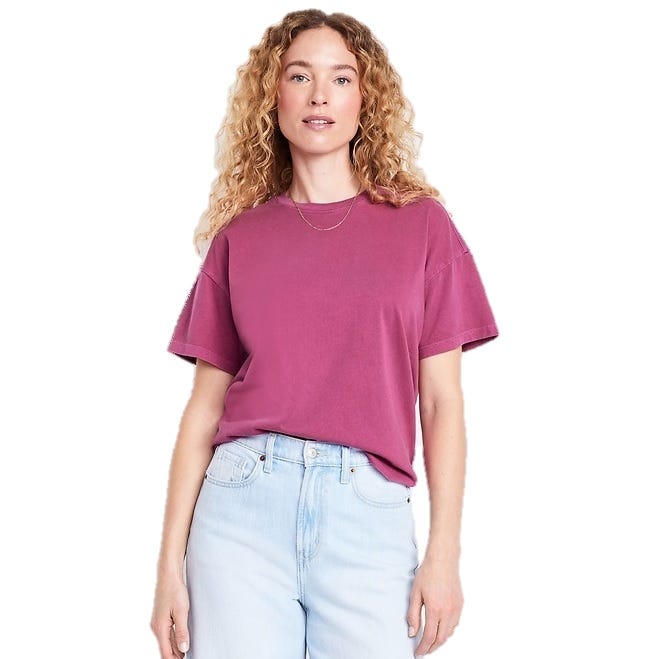 A woman is wearing a pink short-sleeve T-shirt paired with light blue jeans. She has curly hair and is accessorized with a simple necklace.