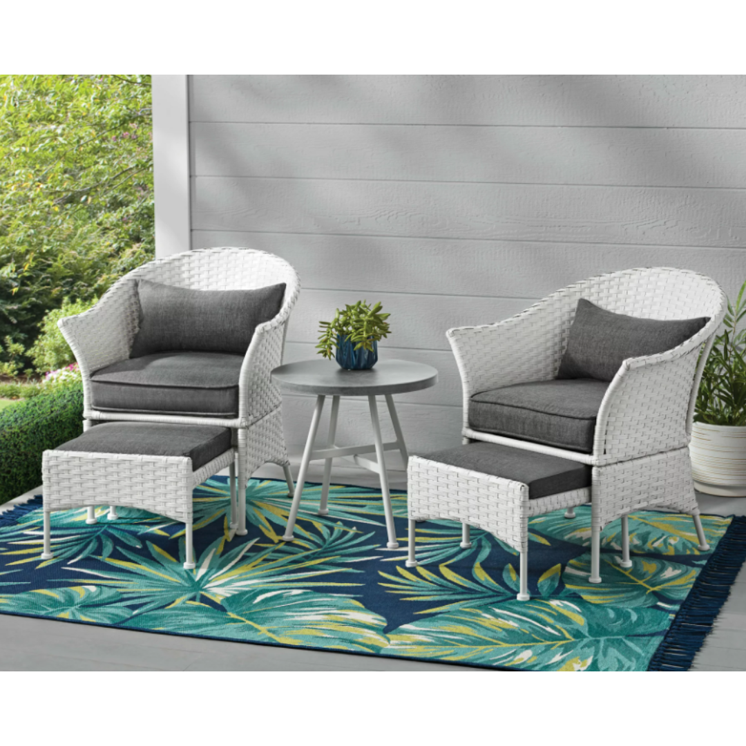 Outdoor furniture set with two white woven chairs with grey cushions, a matching ottoman, and a grey round side table on a leaf-patterned area rug.