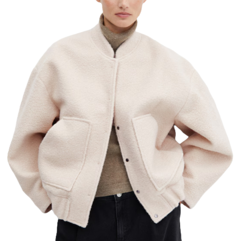 An oversized, soft pink bomber jacket with a large fit, featuring a wooly texture, two front pockets, and snap buttons. It is styled with a high collar and worn over a beige turtleneck sweater.
