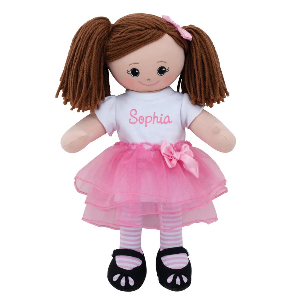A soft doll with brown yarn hair, wearing a white shirt with \“Sophia\“ written on it, a pink tulle skirt, striped leggings, and black shoes.