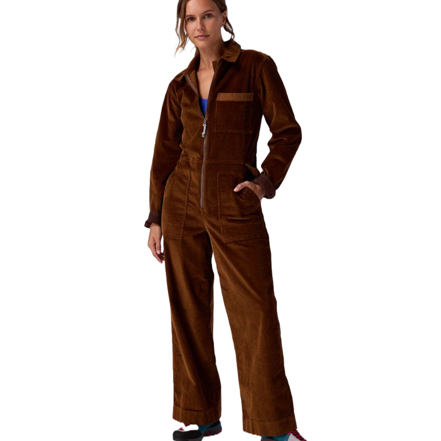 A woman is wearing a brown corduroy jumpsuit featuring a front zipper, long sleeves, wide legs, multiple pocket details, and a collar. She has her hands in the pockets and is wearing sneakers.