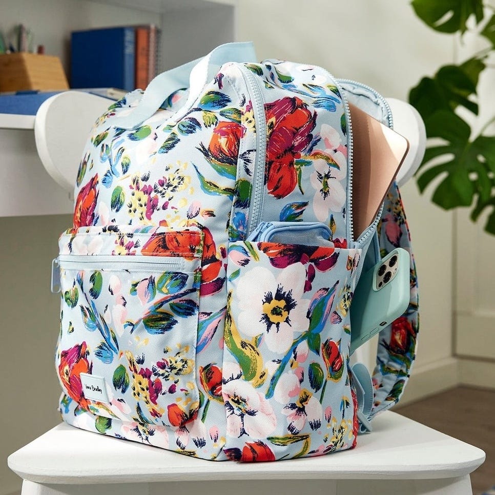 A floral-patterned backpack with a front pocket contains a laptop and a smartphone, resting on a white chair.