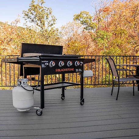 Blackstone outdoor griddle with four burners and a side shelf, next to a propane tank, on a deck with outdoor furniture.