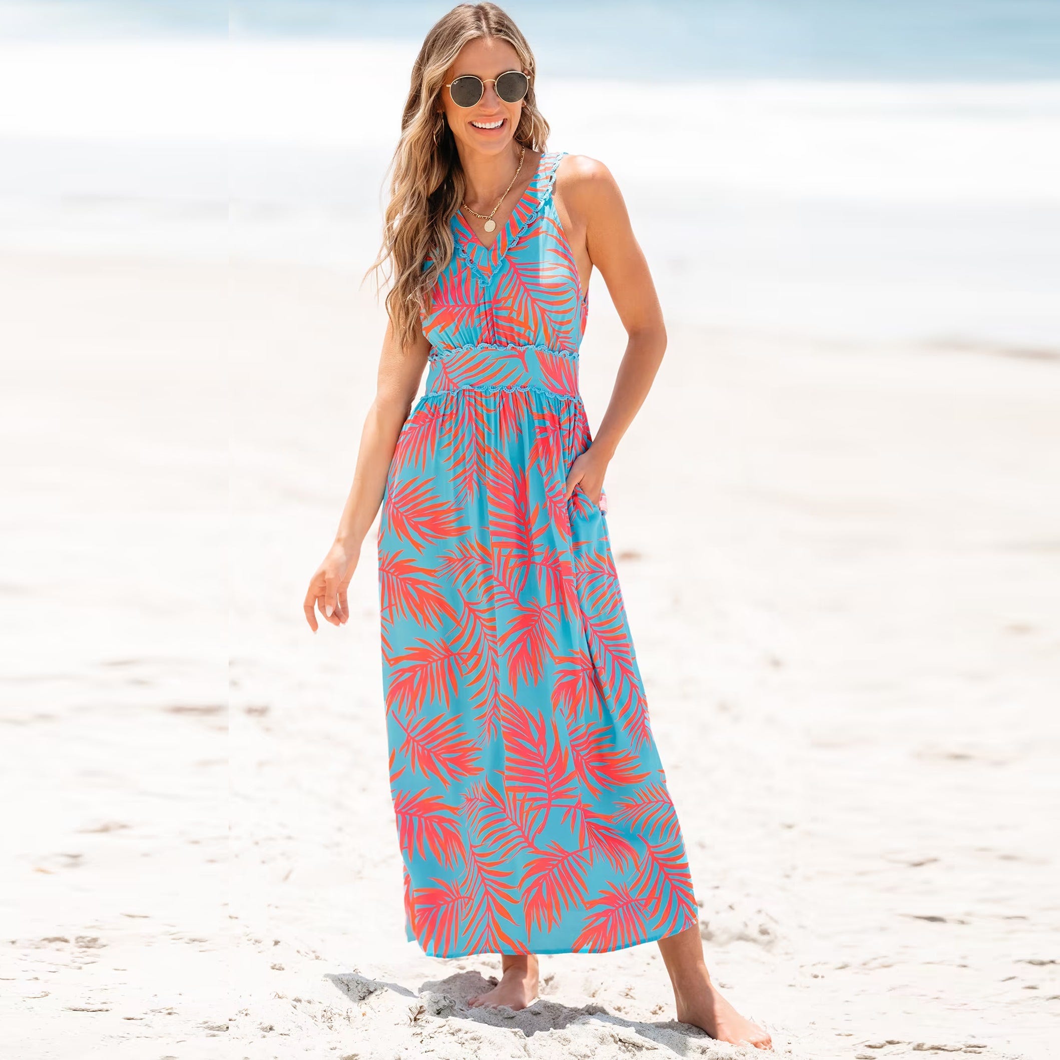 A woman on a beach wearing a sleeveless blue maxi dress with pink and red leaf patterns, paired with round sunglasses.