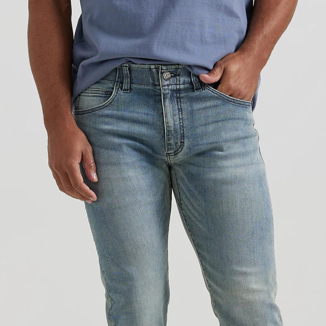 A person is shown wearing light blue, slim-fit Lee Men's Extreme Motion Jeans with a casual blue top tucked partly into the waistband.