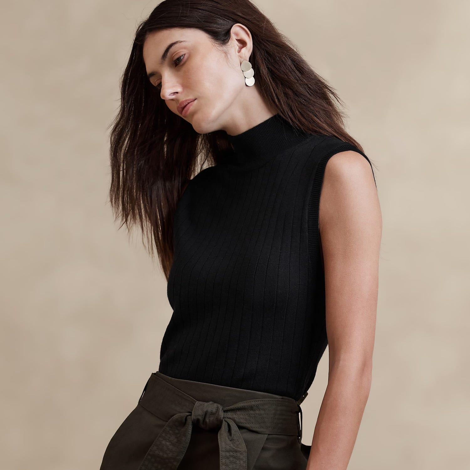 A woman wearing a black turtleneck sleeveless top, olive belt-cinched trousers, and large, circular white earrings.