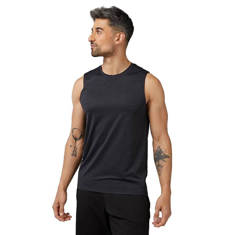 A man wears a sleeveless, dark-colored 32 Degrees Active Tank Top with a crew neck. The top is paired with black shorts, and the man has tattoos on his arms.
