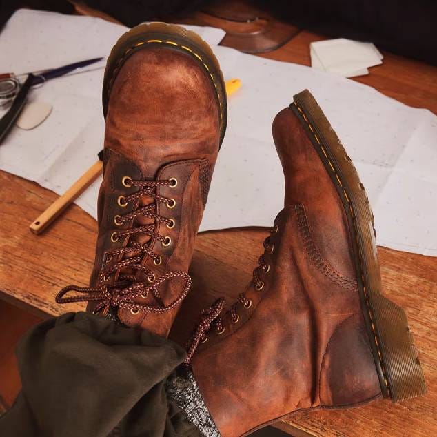 A pair of brown leather lace-up boots with yellow stitching on the soles, next to patterned paper on a wooden surface.