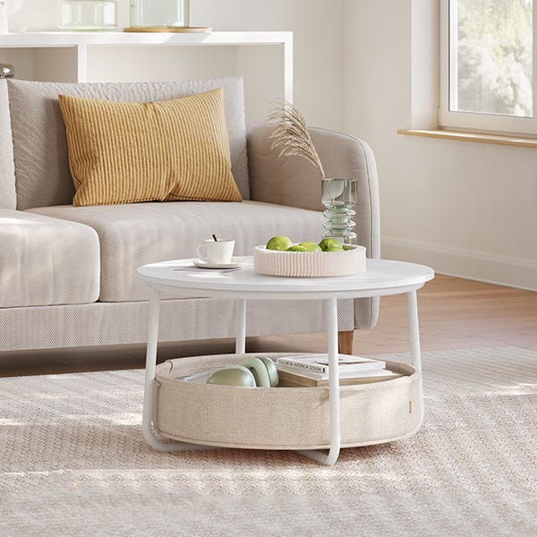 A round white coffee table with a lower shelf holding books and a basket, beside a beige sofa with a yellow pillow.