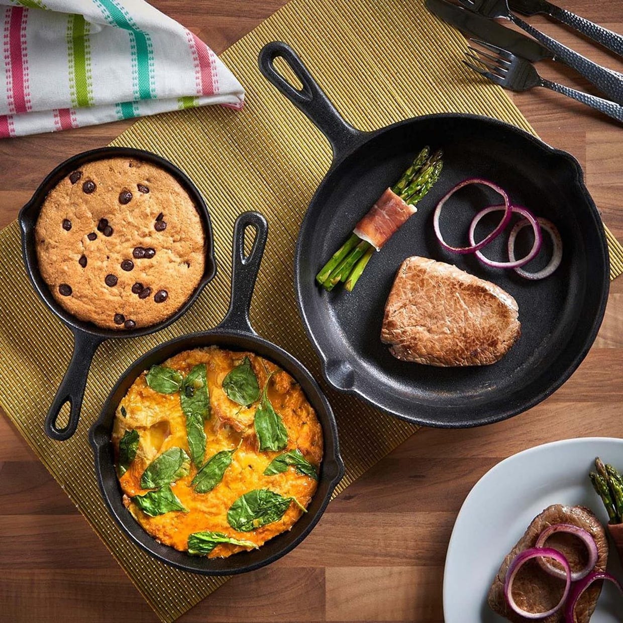 Three cast iron skillets with a chocolate chip cookie, a piece of steak with asparagus and onions, and an egg frittata.