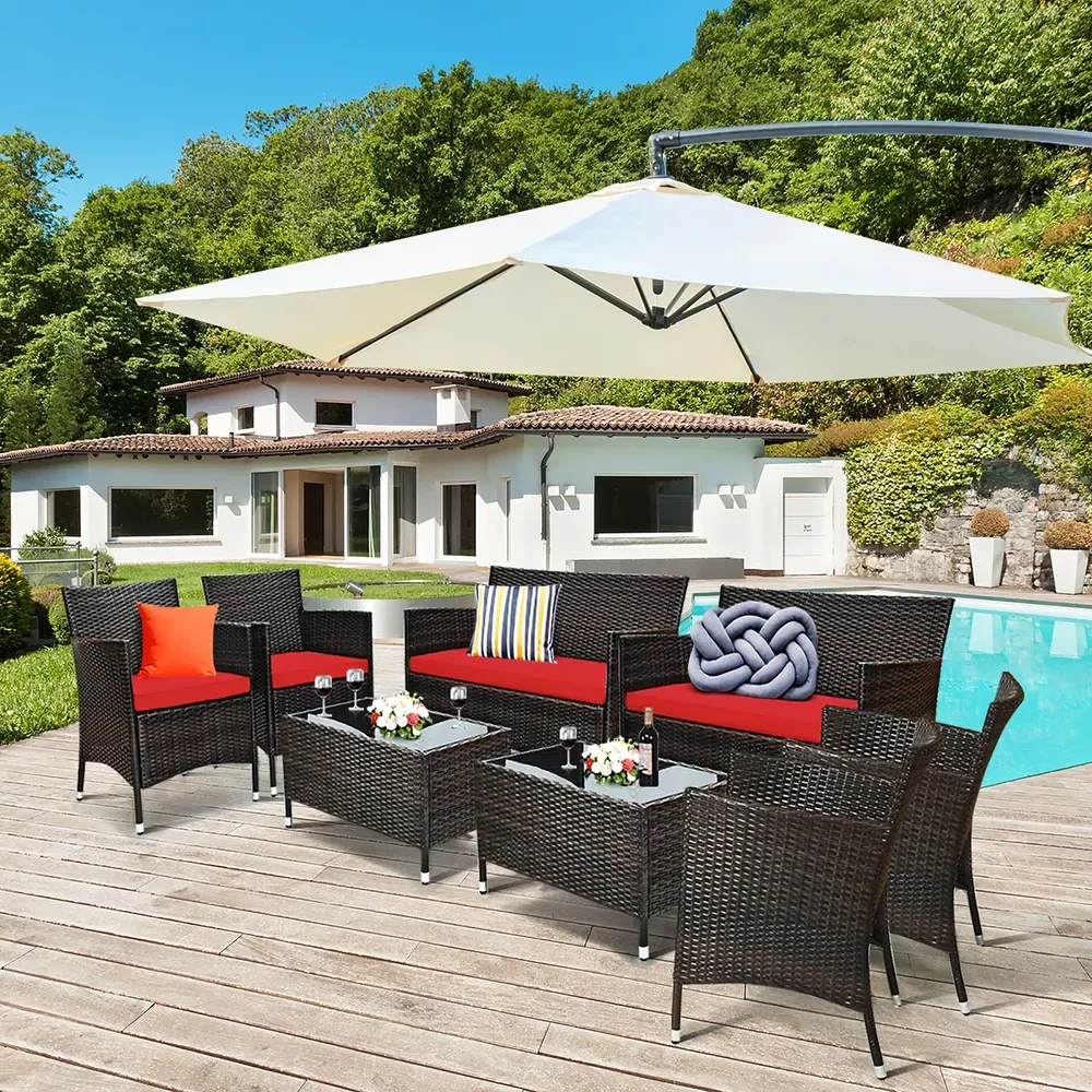 Outdoor patio set with black wicker furniture, including chairs, sofas, and tables, featuring red cushions. A large white umbrella provides shade by a poolside, set against a backdrop of a house and lush greenery.