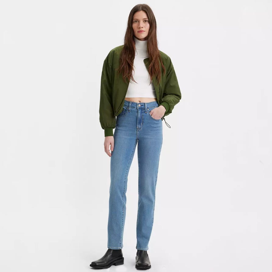 A woman is wearing light blue jeans, a white cropped top, a green bomber jacket, and black boots.