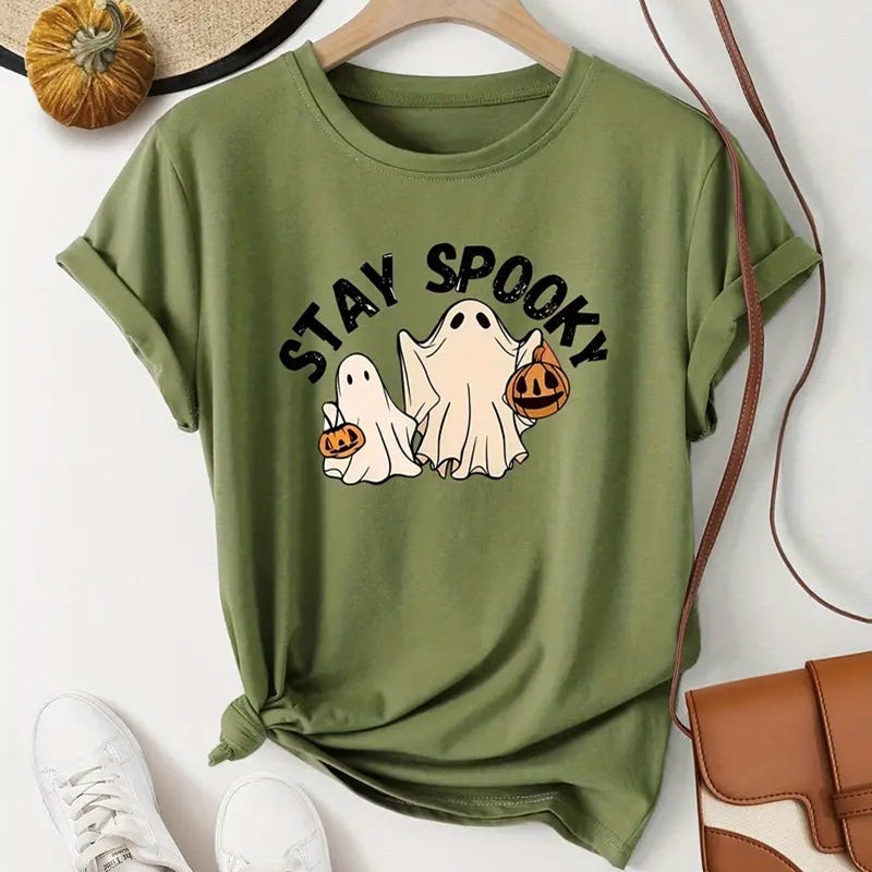 Green t-shirt with a Halloween-themed print featuring cartoon ghosts and the phrase \“STAY SPOOKY.\“ A small pumpkin and white sneakers accompany the shirt.