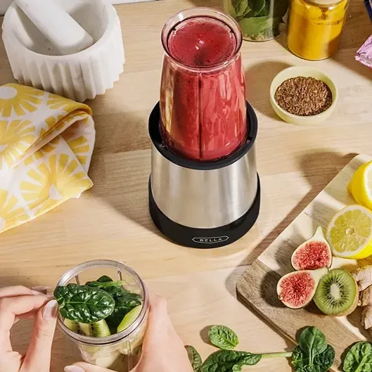 A personal blender with a red smoothie inside and hands preparing ingredients beside it.