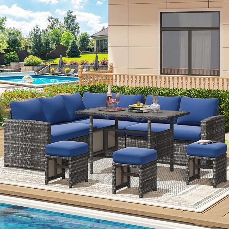 Outdoor patio set featuring blue cushioned seating, a corner sofa, two stools, and a dining table, with a pool and landscaped backyard in the background.