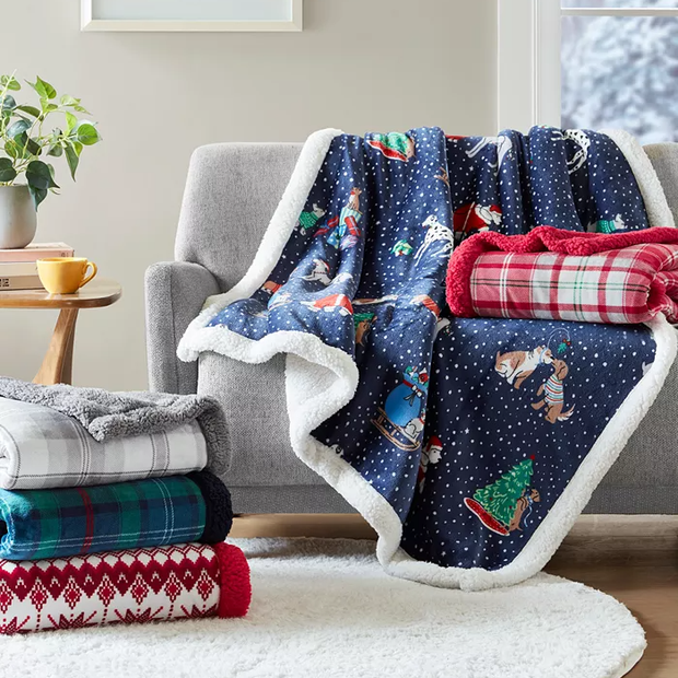 A selection of cozy blankets with various patterns, including a holiday-themed design, is draped over a chair and stacked on the floor.