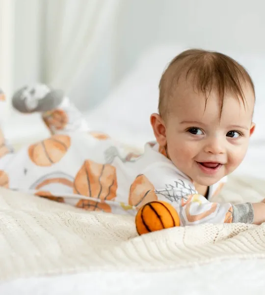 A baby in Burt's Bees Baby pajamas featuring an orange basketball pattern on a white background. The pajamas include long sleeves and footed bottoms for warmth and comfort.