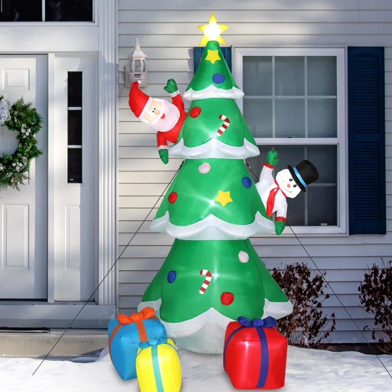 An 8-foot tall inflatable Christmas tree features a snowman and Santa Claus climbing it, with colorful presents at the base. A wreath hangs on the house door behind it.