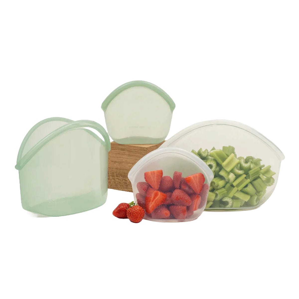 Silicone storage bags in various sizes containing cut strawberries and celery. Several empty bags are also shown, with one resting on a wooden block.
