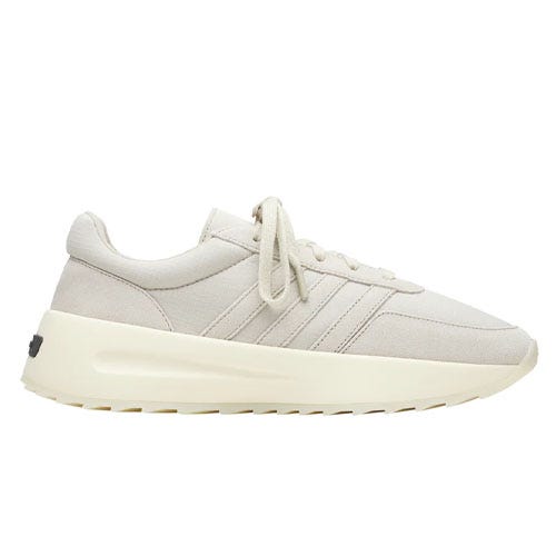 The Adidas x Fear of God sneakers feature a minimalist design in a light beige color with subtle Adidas stripes, a chunky cream sole, and laces for closure, offering a stylish and modern look.