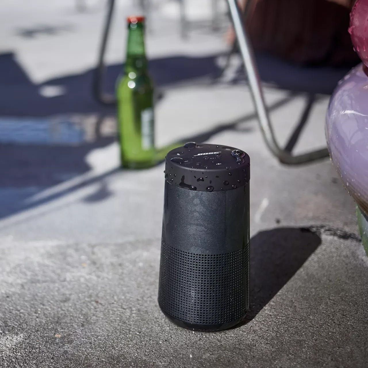 Portable wireless speaker with water-resistance feature, next to a green bottle on a concrete surface.