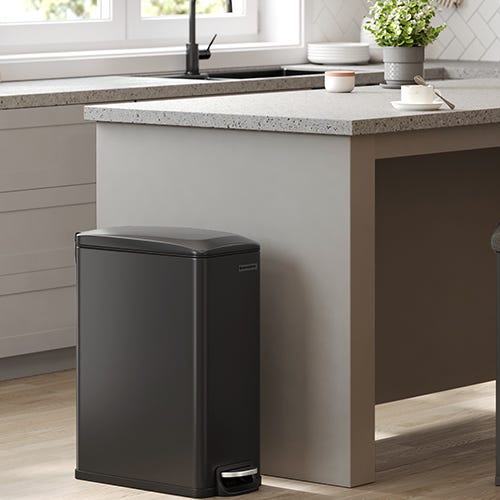 A black, rectangular trash can with a foot pedal stands near a kitchen counter.