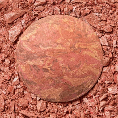 A round makeup compact with a marbled design in shades of pink and brown, surrounded by broken pieces of similar-toned powder.