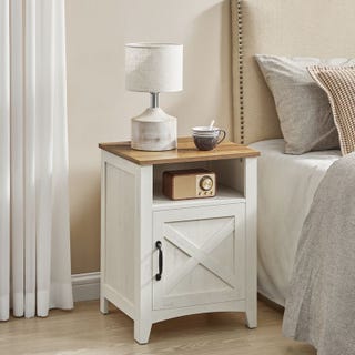A bedside table with a lamp, coffee cup, and vintage radio on top, next to a bed with pillows.