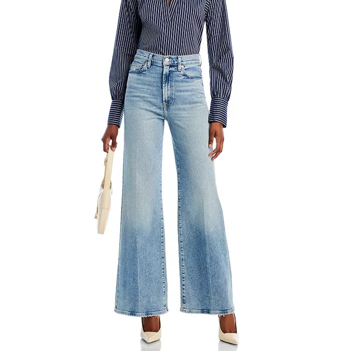 A person is wearing high-waisted, wide-leg jeans in a light blue wash with a subtle fade and front pockets. They are styled with a striped button-up shirt and white heels.