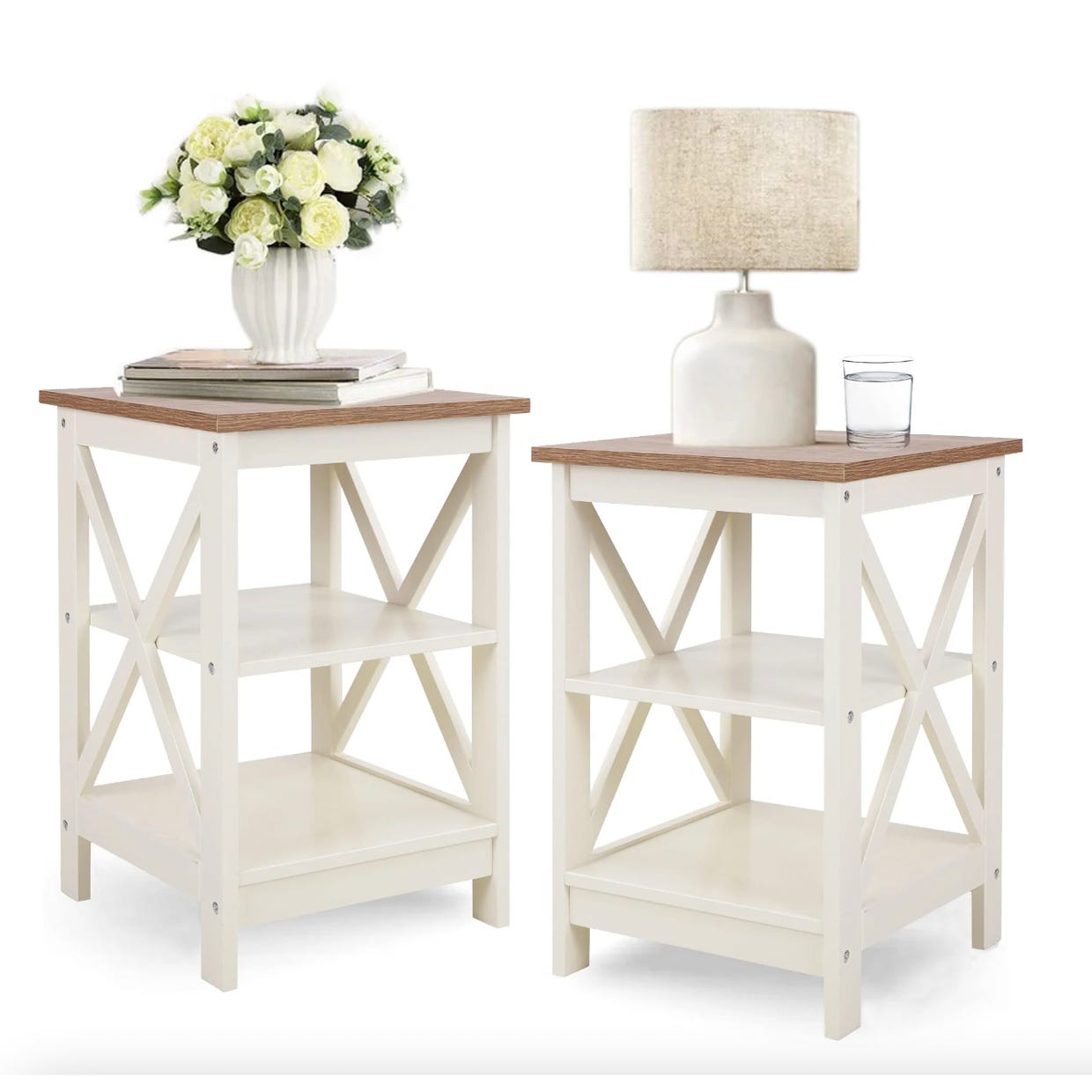 Two white and brown end tables with a cross design on the sides, one with a vase of flowers and one with a lamp and glass.