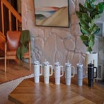 Five white Stanley Straw Tumblers with handles and gray flip straws are displayed on a wooden table, alongside a black tumbler with a handle.