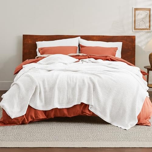 A white waffle weave blanket is draped over a bed with rust-colored bedding and wooden headboard, with pillows matching the sheets.
