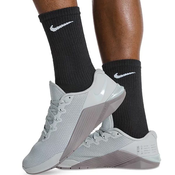 A person wearing light gray Nike Metcon shoes paired with black Nike socks featuring the white Nike swoosh logo.