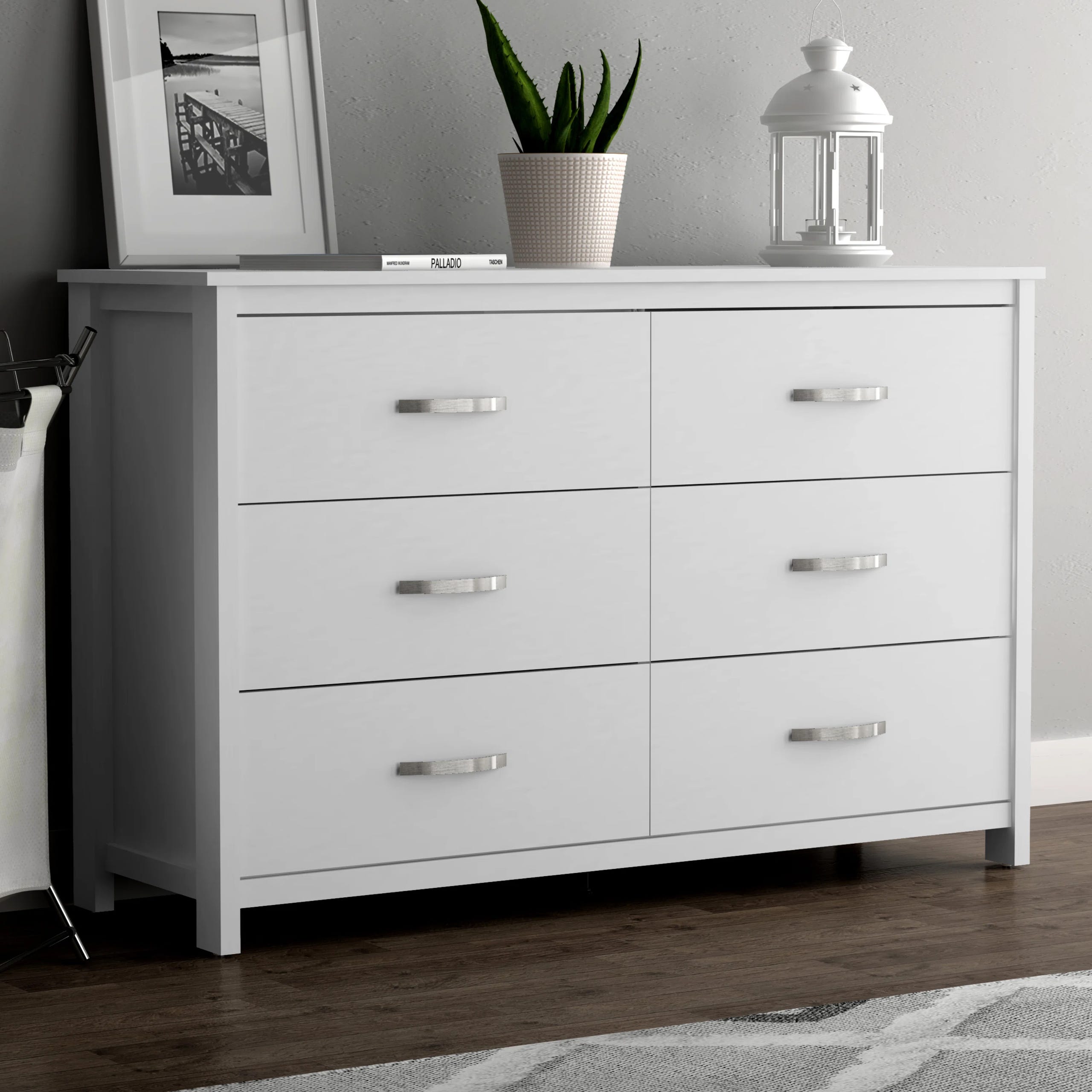 A white six-drawer dresser, with silver handles, accompanied by a potted plant and a lantern on its top.