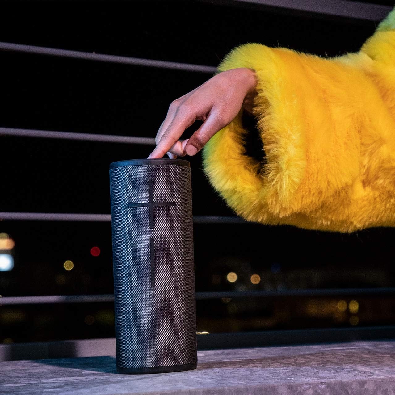 A person in a yellow coat is adjusting a black cylindrical portable speaker on a ledge at night.