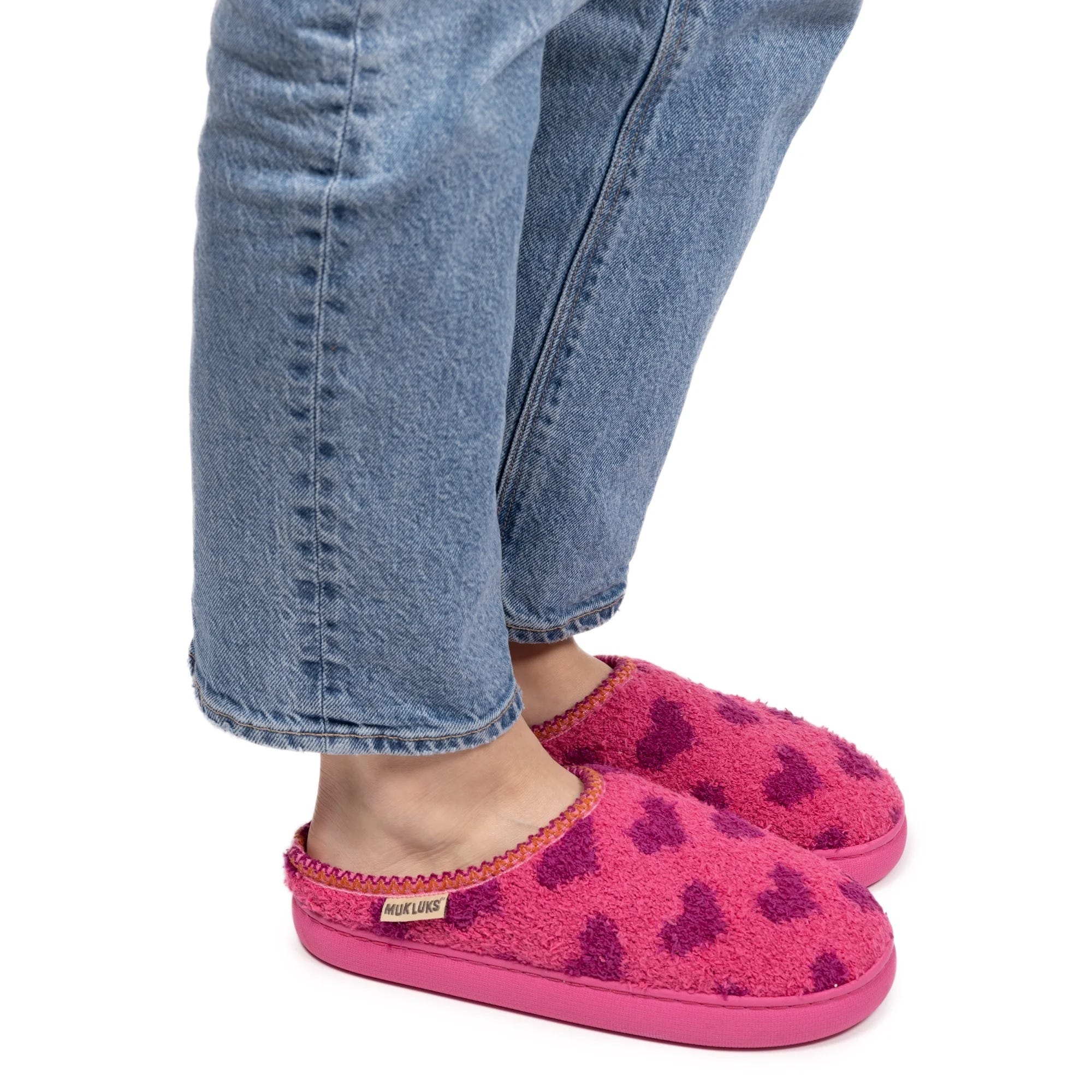 Bright pink Muk Luks slippers with a textured surface, featuring dark pink heart patterns and a contrasting pink sole, worn with cuffed blue jeans.
