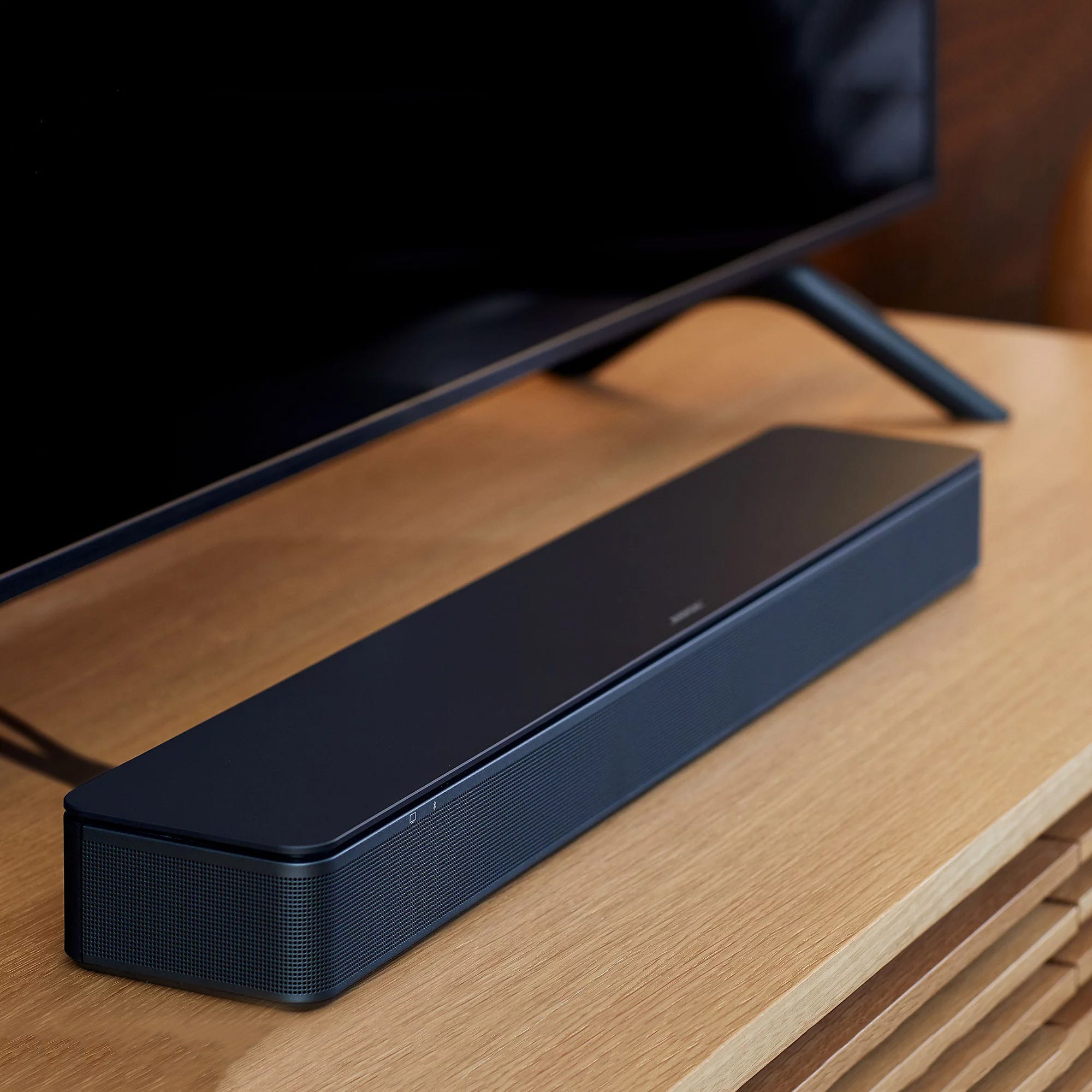 A black soundbar is situated in front of a flat-screen TV on a wooden surface.